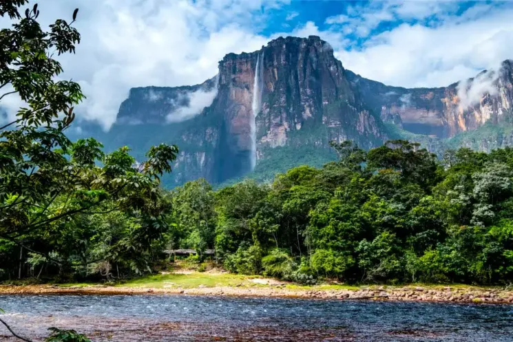 Vista majestosa da cachoeira Salto Ángel despencando de uma imponente montanha de topo plano (tepui) parcialmente envolta em nuvens e névoa. Na base, uma densa floresta tropical verdejante se estende até as margens de um rio de águas calmas que flui em primeiro plano.