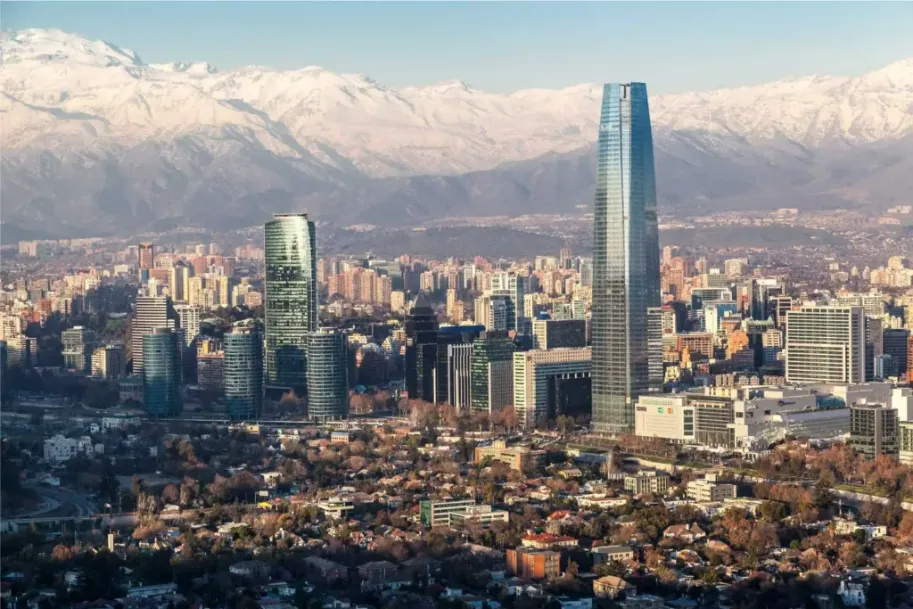 Uma vista panorâmica da cidade de Santiago, Chile, durante um dia claro, com os imponentes Andes cobertos de neve ao fundo. O arranha-céu moderno Costanera Center domina o horizonte.
