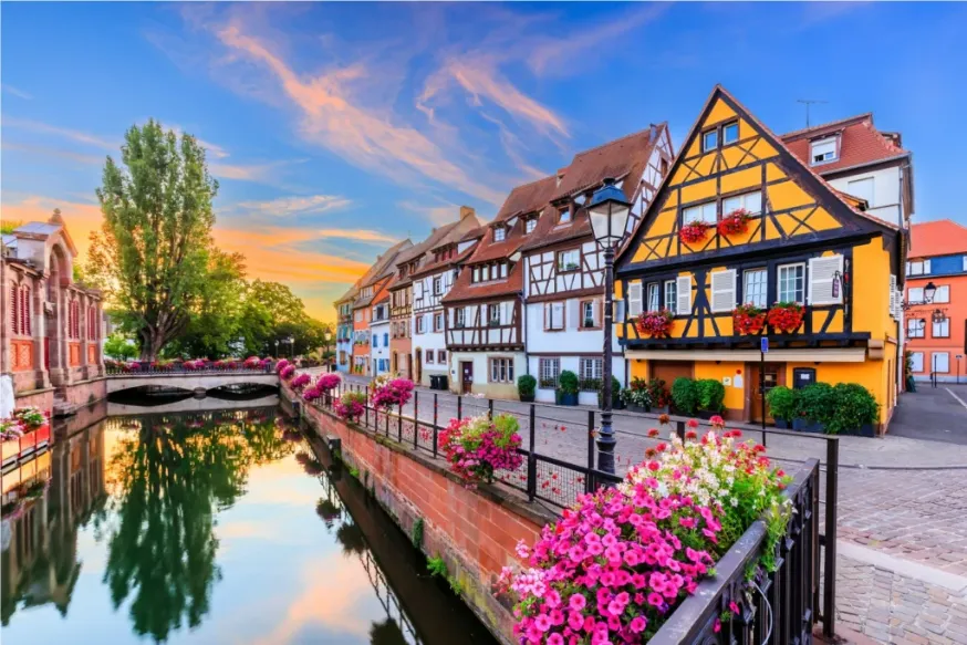 A 'Pequena Veneza' em Colmar, França, ao pôr do sol, com casas de enxaimel coloridas e flores vibrantes refletidas em um canal tranquilo.