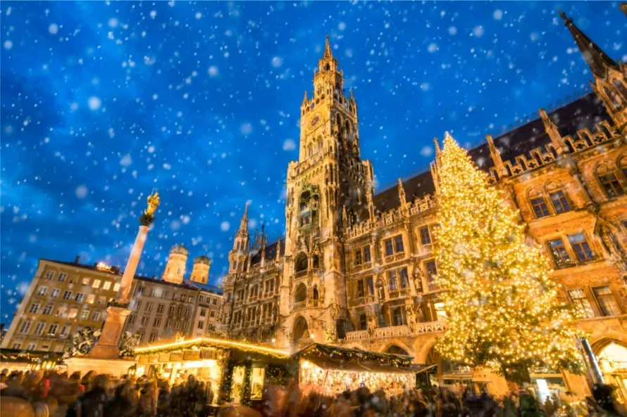 Cena noturna e festiva na praça Marienplatz, em Munique, com neve caindo sob um céu azul escuro. O imponente edifício de arquitetura gótica da Nova Prefeitura (Neues Rathaus) destaca-se ao centro, ladeado por uma grande e brilhante árvore de Natal. Na base da praça, barracas iluminadas de um tradicional mercado de Natal reúnem uma multidão de pessoas.