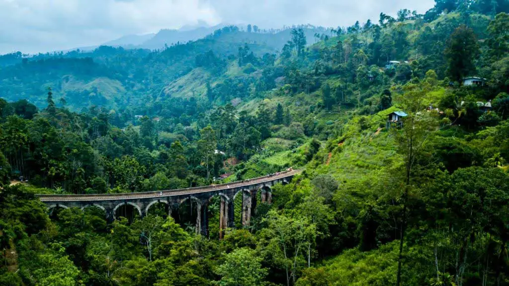 Vista panorâmica de uma majestosa ponte de pedra com múltiplos arcos (conhecida como Ponte dos Nove Arcos) que atravessa um vale profundo cercado por uma densa floresta tropical verdejante. A paisagem montanhosa é coberta por uma vegetação exuberante, com algumas pequenas construções pontilhando as encostas. Ao fundo, montanhas se erguem sob um céu nublado, enquanto pequenas figuras de pessoas podem ser vistas caminhando sobre os trilhos da ponte.