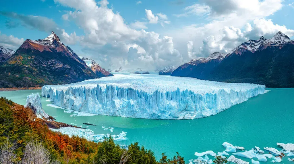 Vista panorâmica do imponente Glaciar Perito Moreno, destacando sua enorme parede de gelo azul e branco que avança sobre um lago de águas azul-turquesa. Vários blocos de gelo flutuam na água ao redor da base. A paisagem é emoldurada por montanhas escuras com picos nevados ao fundo e vegetação com cores de outono em primeiro plano, sob um céu azul com nuvens brancas.