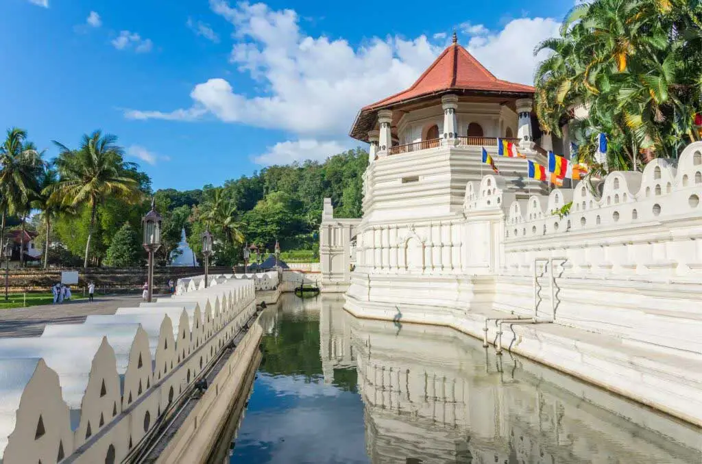 Vista externa do Templo do Dente Sagrado (Sri Dalada Maligawa) em Kandy, no Sri Lanka, em um dia ensolarado. A imagem destaca a estrutura branca do templo com seu característico telhado avermelhado de formato octogonal, adornada com coloridas bandeiras budistas. O complexo é cercado por um tradicional muro branco com recortes ondulados e um fosso de águas calmas, criando um belo reflexo da construção e do céu azul com nuvens brancas. Ao fundo, palmeiras e uma colina coberta por densa vegetação tropical completam a paisagem.