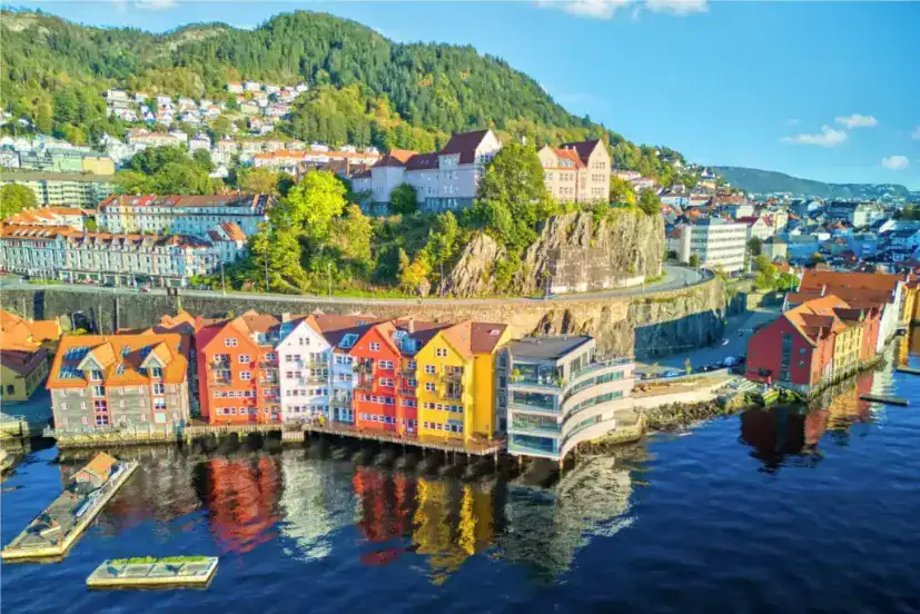Vista aérea da orla colorida de Bergen, Noruega, com montanhas arborizadas ao fundo e reflexos nítidos na água.