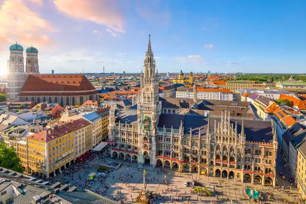 Vista panorâmica da praça Marienplatz em Munique, destacando a imponente Nova Prefeitura (Neues Rathaus) no centro e a igreja Frauenkirche com suas duas torres de cúpulas verdes à esquerda.