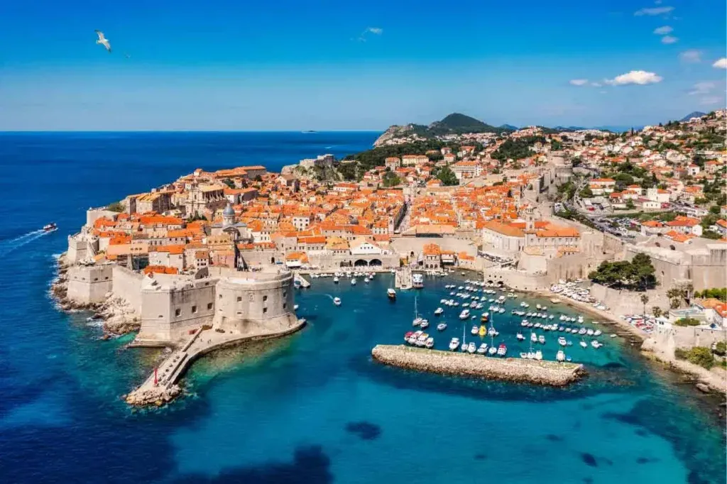 Vista aérea da cidade amuralhada de Dubrovnik, na Croácia, com seus tradicionais telhados de terracota laranja e um porto repleto de barcos cercado pelo mar azul cristalino.