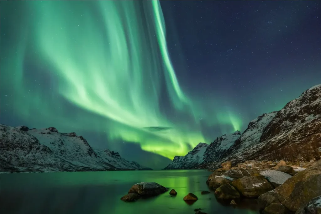 Vibrante aurora boreal verde sobre montanhas nevadas e um fiorde, com reflexos na água e rochas no primeiro plano.