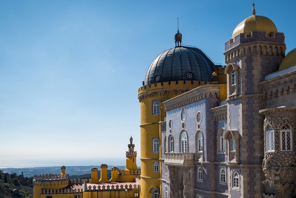 Detalhe arquitetônico do Palácio da Pena em Sintra, Portugal, mostrando as paredes coloridas, as cúpulas ornamentadas e um céu azul claro.
