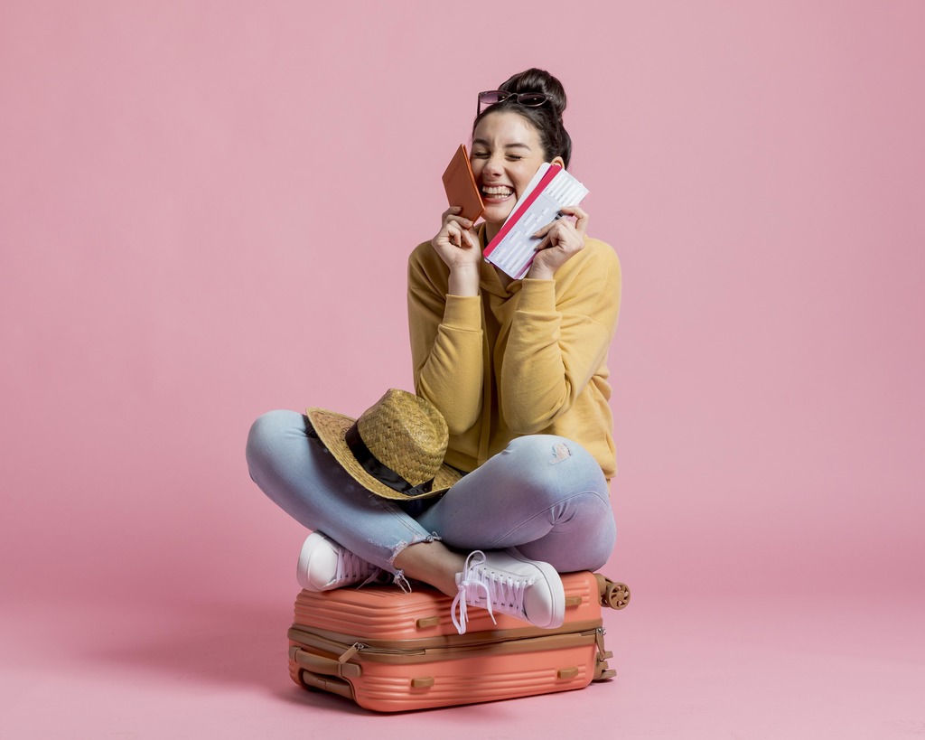 Jovem sentada de perna cruzada sobre uma mala de viagem, com chapéu, livros, e sorrindo ao telefone, contra fundo rosa.