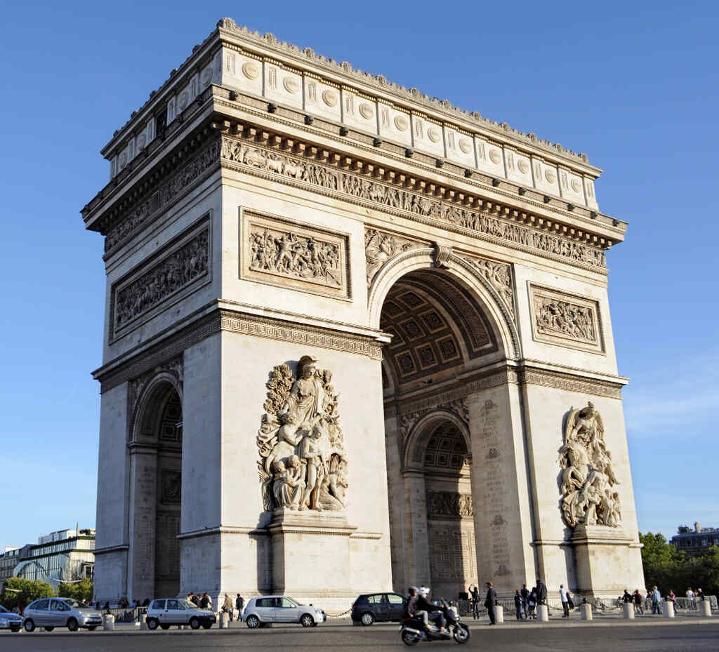 Imagem da Arc de Triomphe em Paris, França, vista de frente, com detalhes das esculturas nas laterais, carros e pessoas ao redor e um céu claro no fundo.