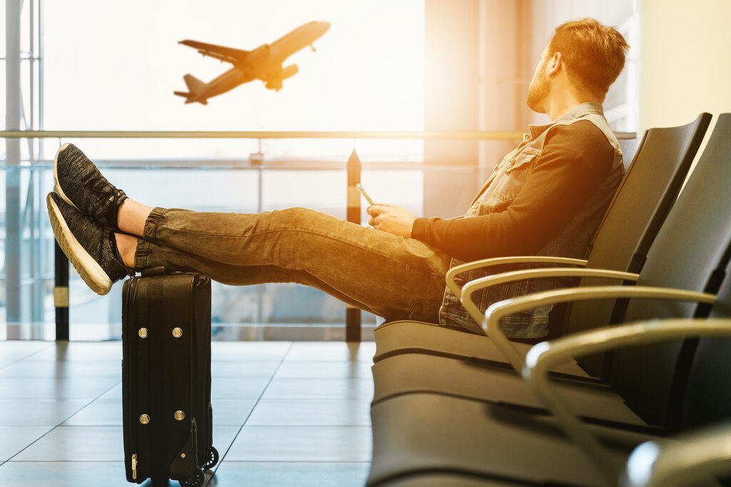 Um homem sentado em uma cadeira de aeroporto com os pés apoiados em uma mala, olhando para um avião decolando ao fundo.