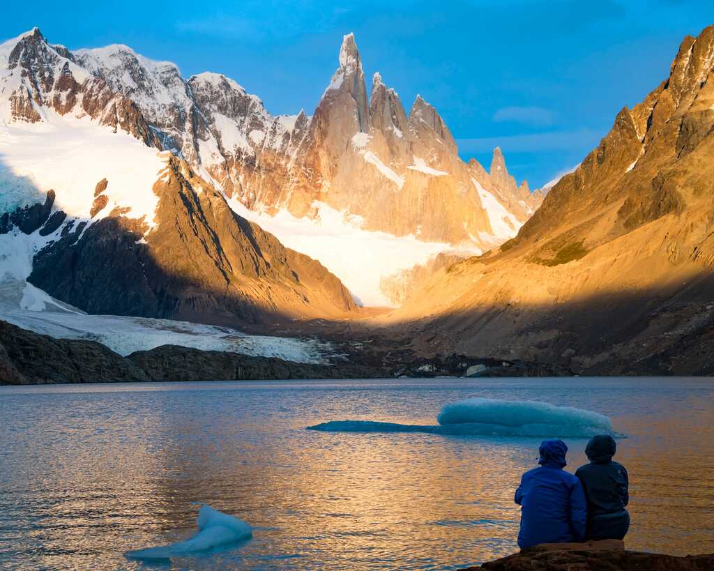 Duas pessoas sentadas à beira de um lago, com montanhas nevadas ao fundo sob um céu azul claro. Icebergs flutuam na água refletindo a luz dourada do sol.