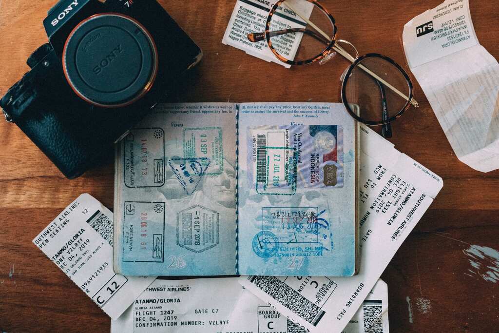 Uma foto de uma mesa com uma câmera, passaporte, óculos e bilhetes de embarque. O passaporte está aberto, mostrando vários selos de visto.