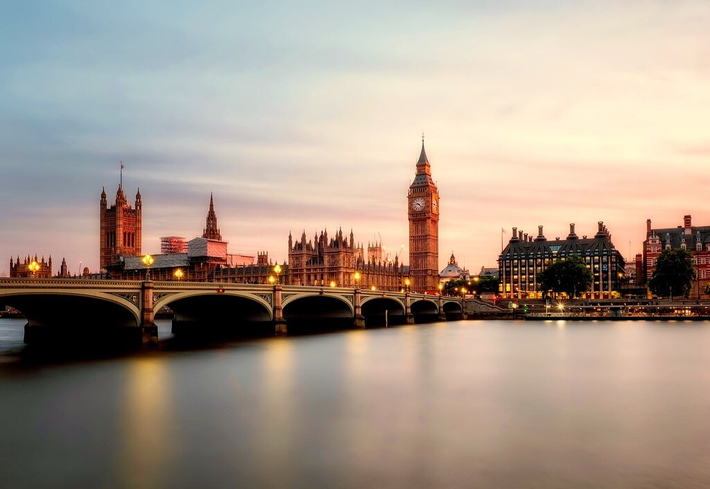 Uma vista aérea panorâmica de Londres, destacando o rio Tâmisa, as Casas do Parlamento e o Big Ben sob um céu dramático com nuvens e raios de sol dourados.
