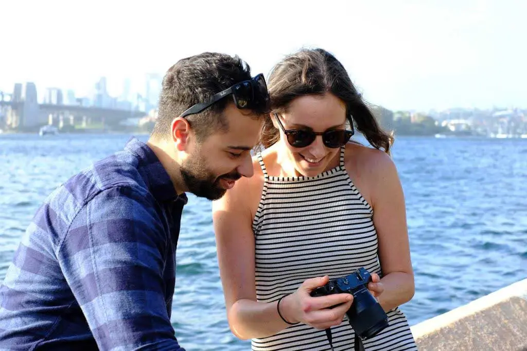 Um close de um casal tirando fotos à beira-mar, ambos sorrindo e se concentrando na tela da câmera.