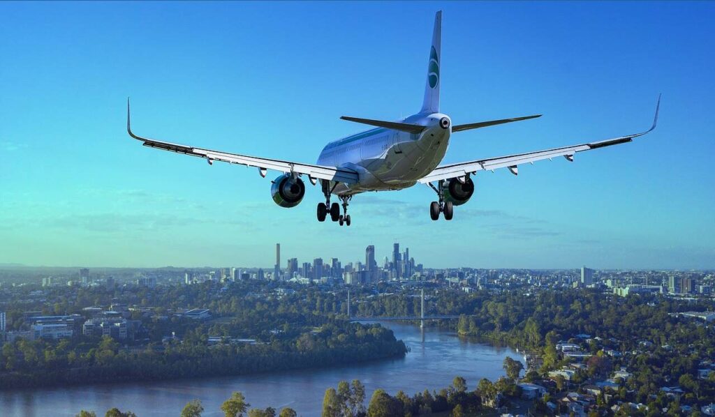 Um avião comercial voando sobre uma cidade com um rio e uma linha do horizonte visível.