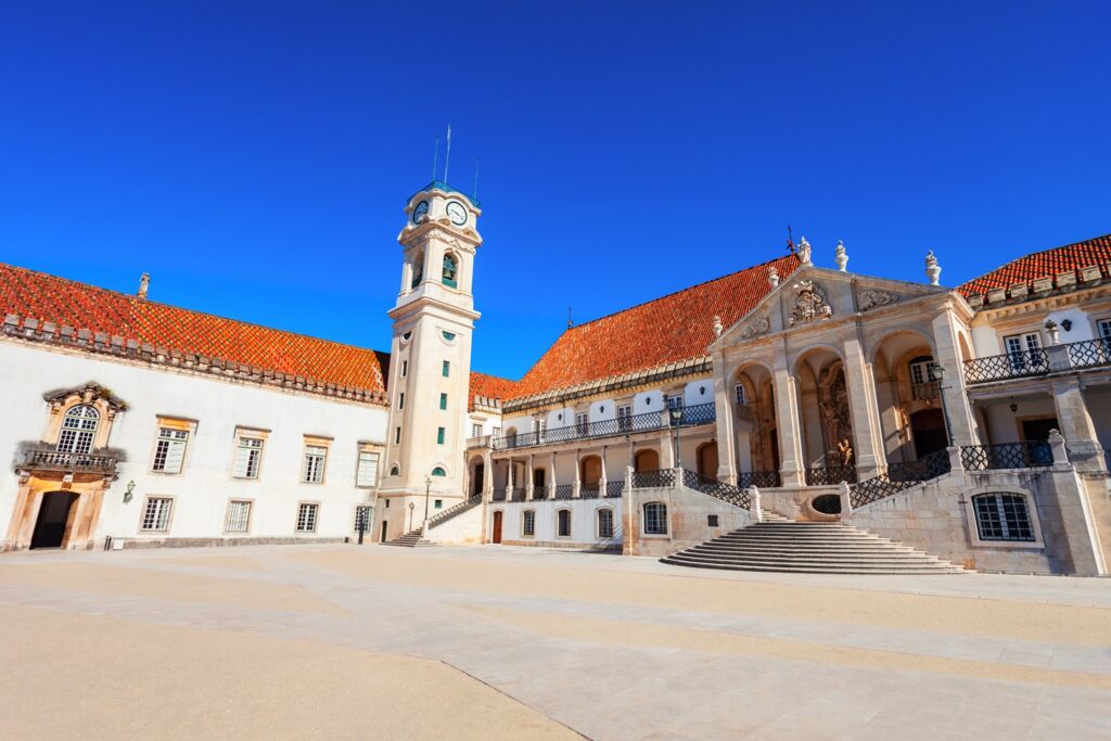 Um grande edifício histórico com fachada branca e telhados vermelhos. À esquerda, há uma torre do relógio e, ao centro, uma escadaria que leva a um pórtico com colunas ornamentadas. O céu está azul e claro, realçando os detalhes arquitetônicos do prédio.
