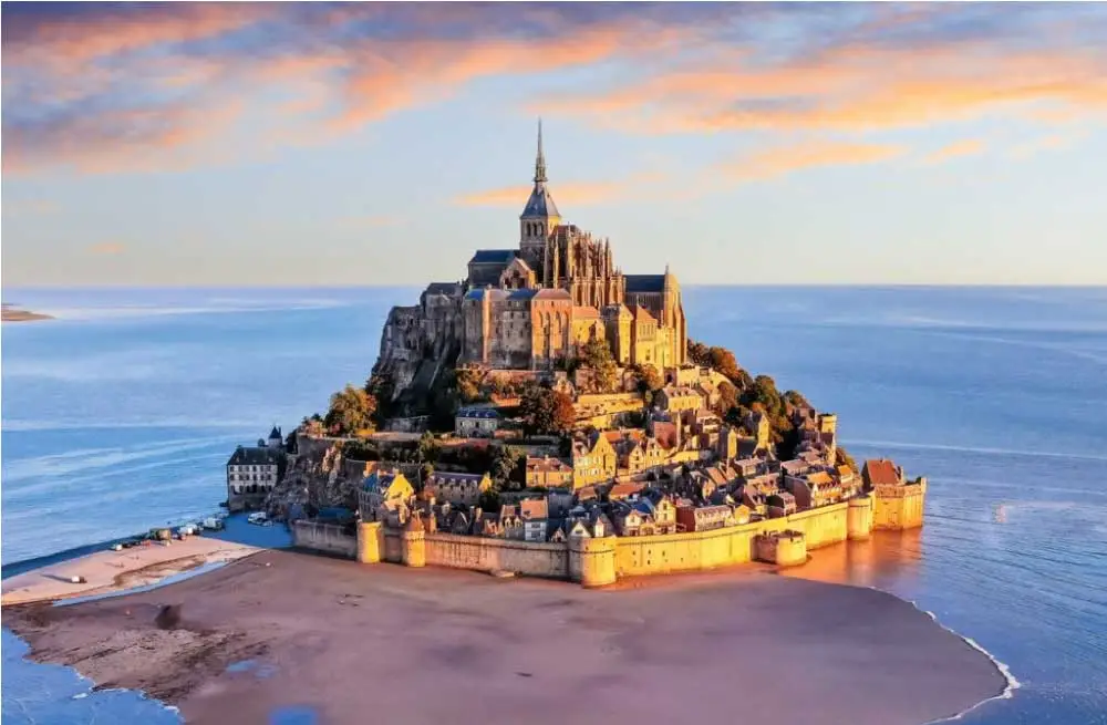 Vista de Mont Saint-Michel, uma ilha com uma abadia medieval, cercada por água e areia sob um céu de pôr do sol.