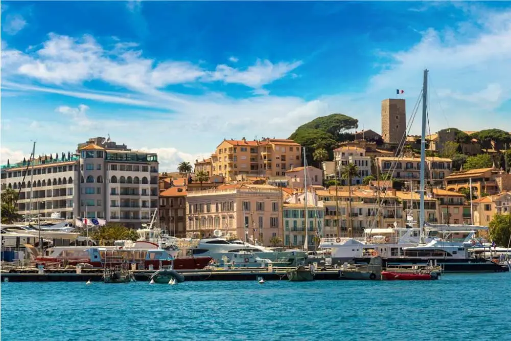 Vista do porto de Cannes, com edifícios históricos na encosta, iates luxuosos e céu azul com nuvens.