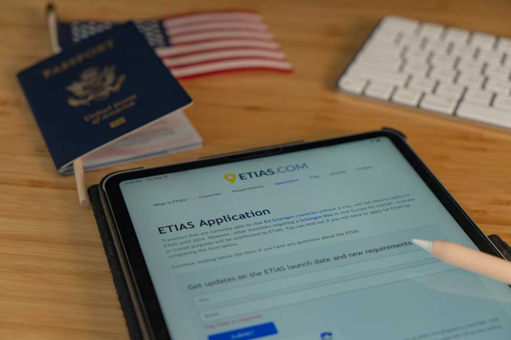 Sobre uma mesa, detalhe de um formulário ETIAS, acompanhado de um passaporte, uma bandeira, uma caneta e um teclado.