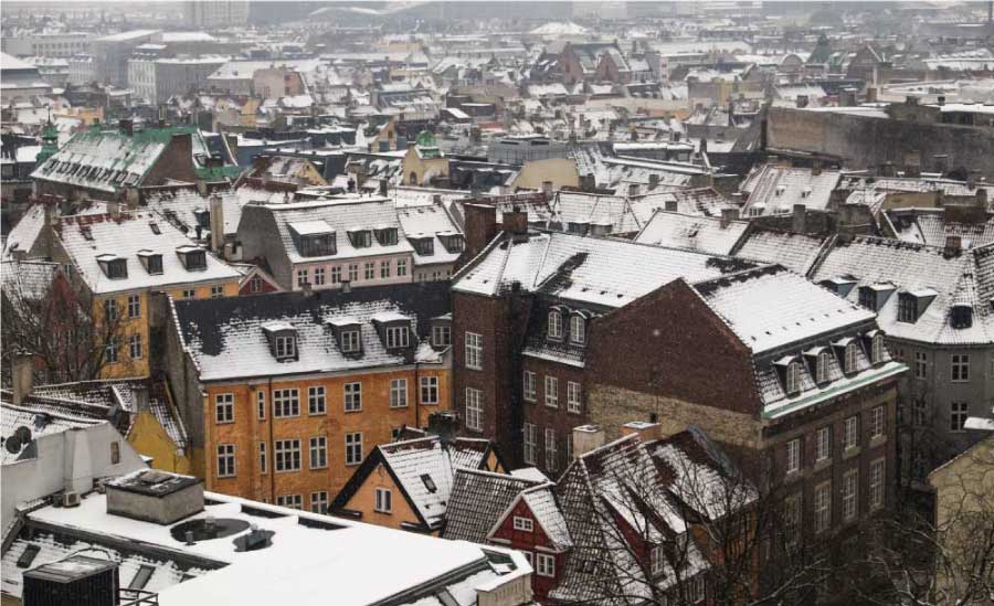 Vista aérea de uma cidade com telhados de casas cobertos por neve, destacando prédios de arquitetura europeia em tons de amarelo, vermelho e tijolos, em um dia de inverno.