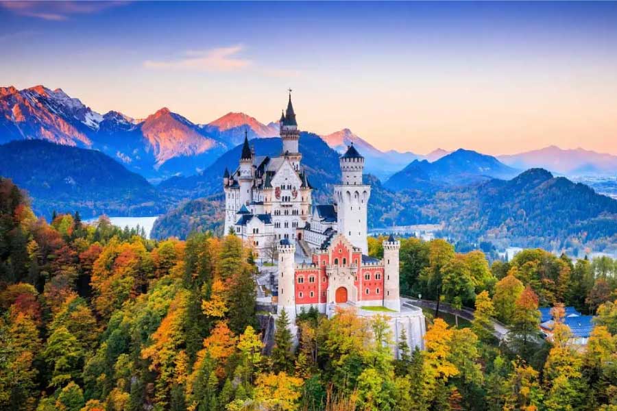 Um castelo majestoso de conto de fadas localizado no topo de uma colina cercada por árvores com folhas de outono, que variam em tons de verde, laranja e amarelo. Ao fundo, há montanhas com picos cobertos de neve, iluminados pela luz suave do entardecer, e um céu gradiente com tons de azul e rosa.