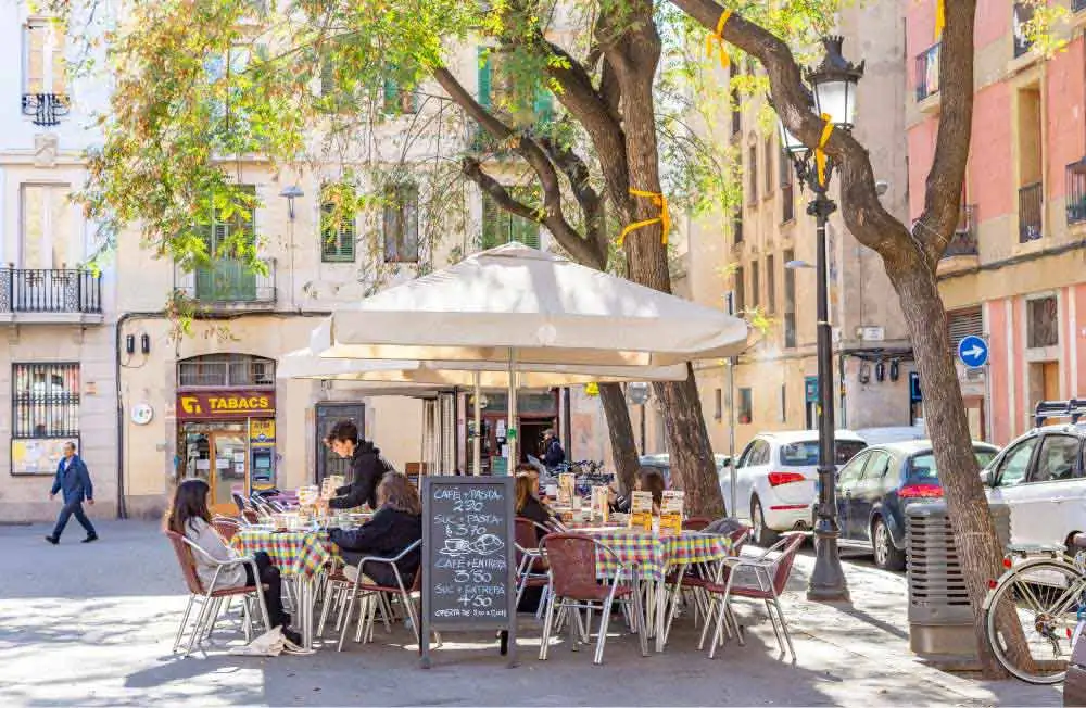 Uma cena de rua com um café. Mesas com toalhas de mesa xadrez estão montadas sob guarda-sóis. Pessoas estão sentadas às mesas. Uma placa de menu é visível. Edifícios ladeiam a rua e árvores são intercaladas.