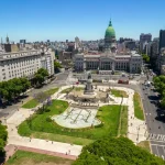 Uma fotografia aérea do Congresso da Nação Argentina, localizado em Buenos Aires. O prédio proeminente está no centro da imagem, rodeado por edifícios da cidade e uma praça verde. O céu é azul claro, com algumas nuvens visíveis.
