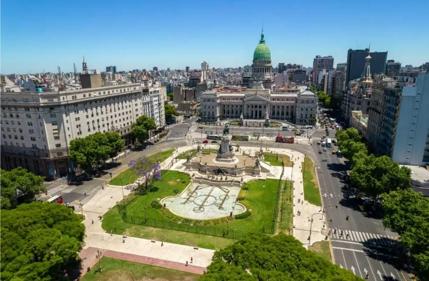 Uma fotografia aérea do Congresso da Nação Argentina, localizado em Buenos Aires. O prédio proeminente está no centro da imagem, rodeado por edifícios da cidade e uma praça verde. O céu é azul claro, com algumas nuvens visíveis.