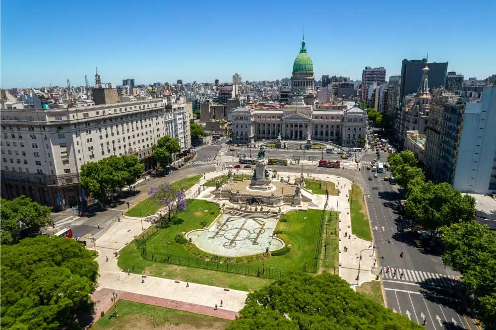 Uma fotografia aérea do Congresso da Nação Argentina, localizado em Buenos Aires. O prédio proeminente está no centro da imagem, rodeado por edifícios da cidade e uma praça verde. O céu é azul claro, com algumas nuvens visíveis.
