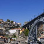 Uma vista panorâmica da Ponte Dom Luís I na cidade do Porto, Portugal, em um dia ensolarado, com casas e edifícios empilhados na colina ao fundo.