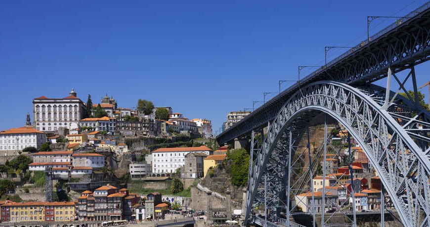 Uma vista panorâmica da Ponte Dom Luís I na cidade do Porto, Portugal, em um dia ensolarado, com casas e edifícios empilhados na colina ao fundo.