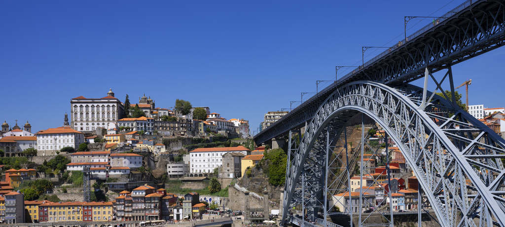Uma vista panorâmica da Ponte Dom Luís I na cidade do Porto, Portugal, em um dia ensolarado, com casas e edifícios empilhados na colina ao fundo.