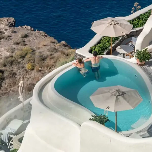 Vista aérea de uma piscina azul cercada por edifícios brancos e penhascos rochosos com vista para o oceano.