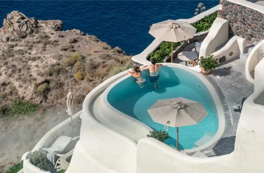 Vista aérea de uma piscina azul cercada por edifícios brancos e penhascos rochosos com vista para o oceano.