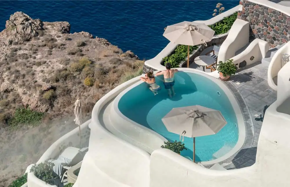 Vista aérea de uma piscina azul cercada por edifícios brancos e penhascos rochosos com vista para o oceano.