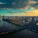 Uma fotografia pitoresca da Ponte de Westminster e das Casas do Parlamento em Londres ao pôr do sol, com o rio Tâmisa calmo em primeiro plano.