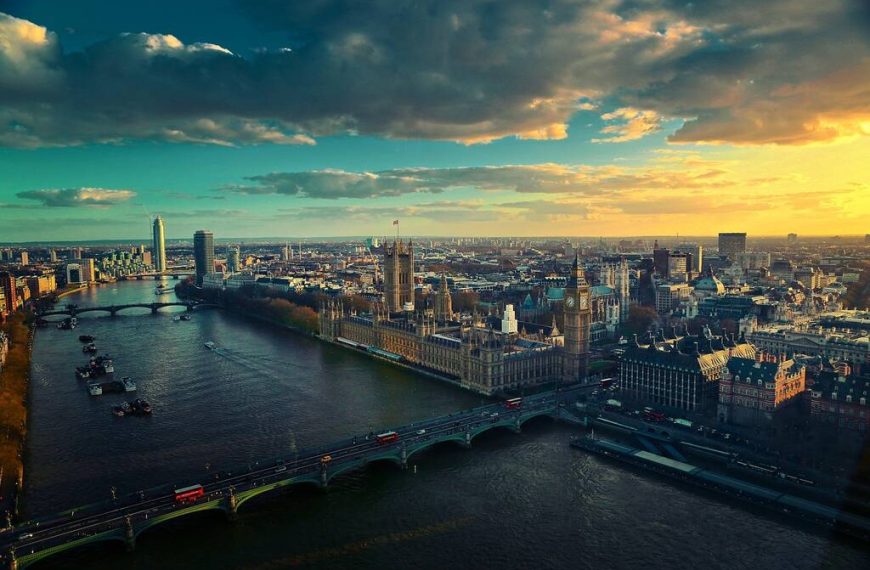 Uma fotografia pitoresca da Ponte de Westminster e das Casas do Parlamento em Londres ao pôr do sol, com o rio Tâmisa calmo em primeiro plano.