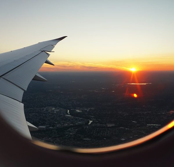 Uma foto tirada de dentro de um avião, mostrando a asa e uma janela com uma vista de um pôr do sol laranja e dourado sobre uma cidade abaixo.