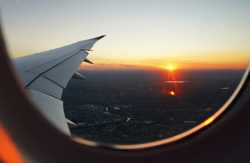Uma foto tirada de dentro de um avião, mostrando a asa e uma janela com uma vista de um pôr do sol laranja e dourado sobre uma cidade abaixo.