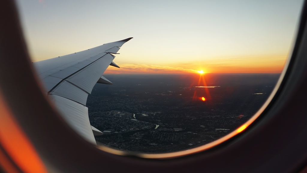 Uma foto tirada de dentro de um avião, mostrando a asa e uma janela com uma vista de um pôr do sol laranja e dourado sobre uma cidade abaixo.
