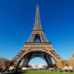 Fotografia da Torre Eiffel a partir do solo, mostrando sua estrutura de treliça de ferro intrincada com o céu azul vívido ao fundo.