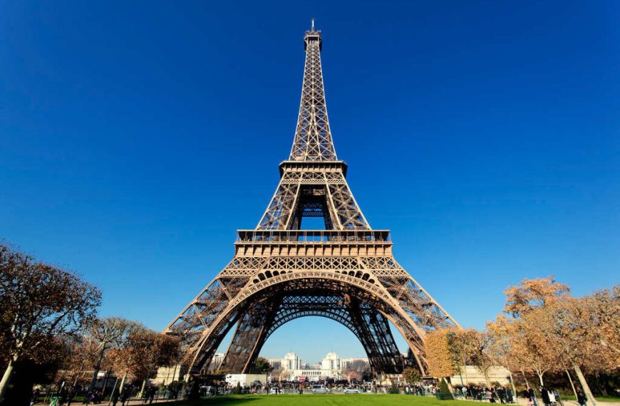 Fotografia da Torre Eiffel a partir do solo, mostrando sua estrutura de treliça de ferro intrincada com o céu azul vívido ao fundo.