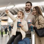 Família de três pessoas felizes no aeroporto, com a criança segurando passagens, todos sorrindo e comemorando a viagem.