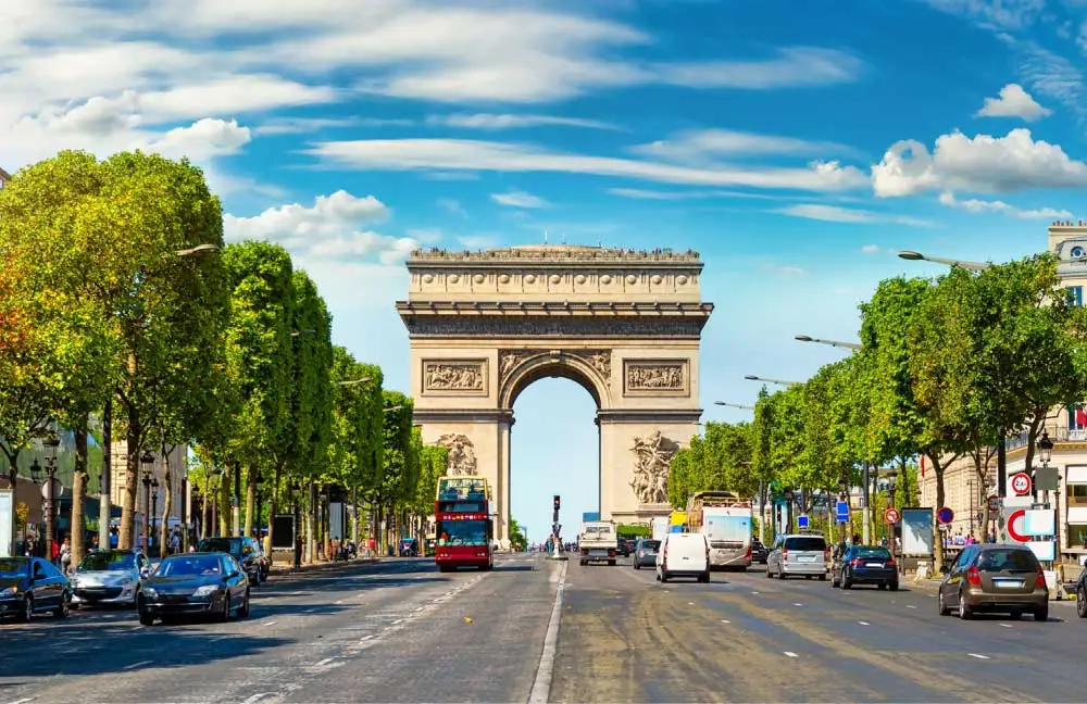 O Arco do Triunfo em Paris, visto de uma rua movimentada alinhada com árvores.