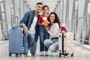 Família de três posando com bagagem em um terminal de aeroporto. A filha está segurando um passaporte e um bilhete.
