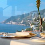 Mesa posta com taças de vidro junto a uma janela, vista panorâmica do mar e da costa ao fundo.