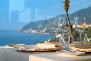 Mesa posta com taças de vidro junto a uma janela, vista panorâmica do mar e da costa ao fundo.