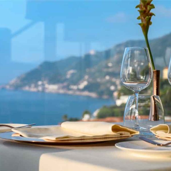 Mesa posta com taças de vidro junto a uma janela, vista panorâmica do mar e da costa ao fundo.