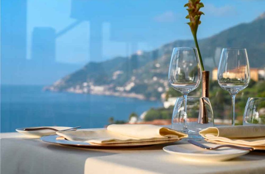 Mesa posta com taças de vidro junto a uma janela, vista panorâmica do mar e da costa ao fundo.